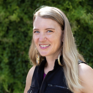 This is a headshot of Anne Heintz. She has light skin and long, straight blond hair. She has blue eyes and is smiling and facing the camera. She is wearing a black vest, gold hoop earrings and is in front of somewhat blurry green foliage in the background.