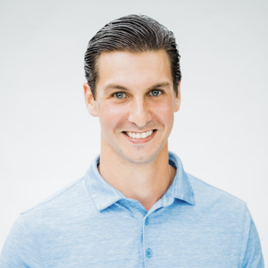 This is a headshot of Zach Rondot. Zach is smiling and facing the camera. He has short brown hair, light skin, and blue eyes. He is wearing a blue collared, button down shirt. The background is light grey and nondescript.