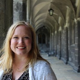 This is a headshot of Alison Keller. Alison has light skin and straight blond hair that galls past her shoulders. Alison is smiling and facing the camera while wearing a dark shirt with a white cardigan over it. The background is a hallway with an arched ceiling and grey brick walls.