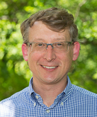 This is a headshot of Cary Roseth. He is smiling and facing the camera. He has light skin and glasses. He has short hair that is medium brown. He is wearing a checkered, blue shirt. The background is slightly blurry foliage.