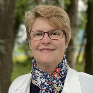 This is a headshot of Colleen Farris. Colleen is facing the camera with a slight smile. Colleen has short, blond hair; light skin, and red-ish glasses. Colleen is wearing a white jacket, black shirt, and colorful floral scarf. The background is trees and foliage that are just out of focus.