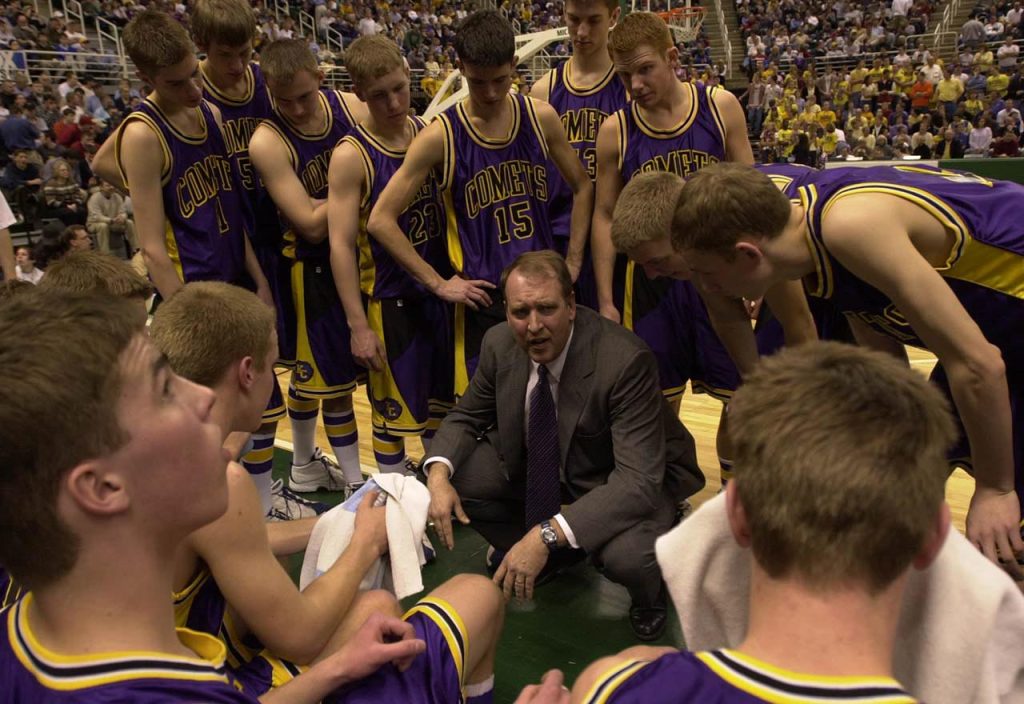 Coach kneels on floor of basketball court and talks strategy with players