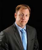 Andrew Nay headshot. Andrew wears a light blue button down shirt and a complementary tie with a black blazer. Andrew's hair is cropped short. 