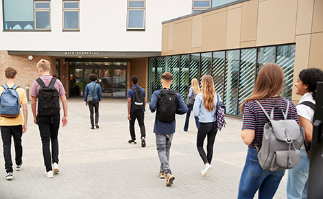 Several students walk toward the door to a school building, wearing backpacks. 