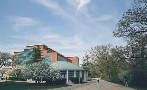 Erickson Hall in spring. The five-story building is comprised of a reddish brick and many horizontal windows. At the forefront of the frame and building, a circular room known as the Kiva is shown.