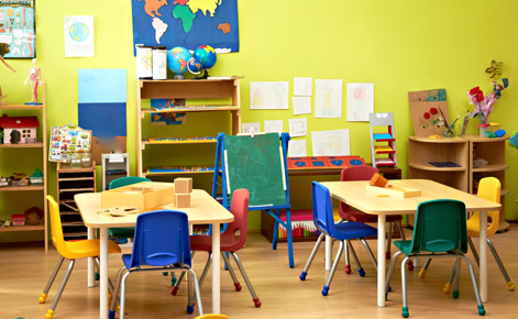 Preschool classroom with yellow walls and multicolored objects in forefront and background, including art supplies, chalkboard easels and tables and chairs.