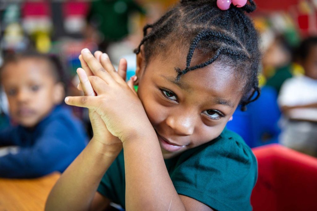 A second grade female student rests her arms on her desk while looking at camera.