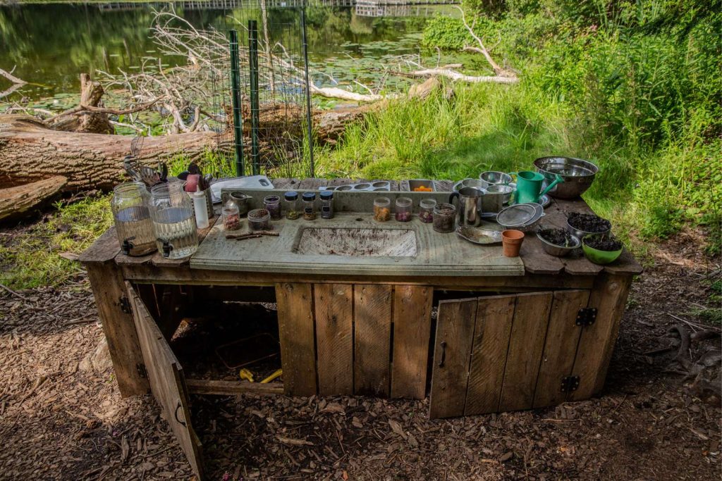 An old wooden countertop with shelving underneath sits outdoors. There are jars and pitchers filled with water on top of the countertop.