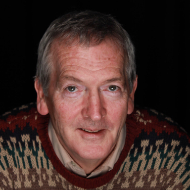 This is a headshot of Chris. He has light skin and greying hair. He is smiling at the camera and wearing a multicolored sweater. 