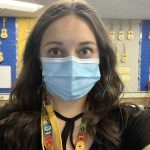 This is a picture of Emily. She has brown hair and is wearing a mask while in her classroom. There are ukuleles hanging on the wall in the background.