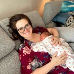 This is a picture of Kathryn. She has brown hair, wears glasses, and is smiling while holding a baby.o