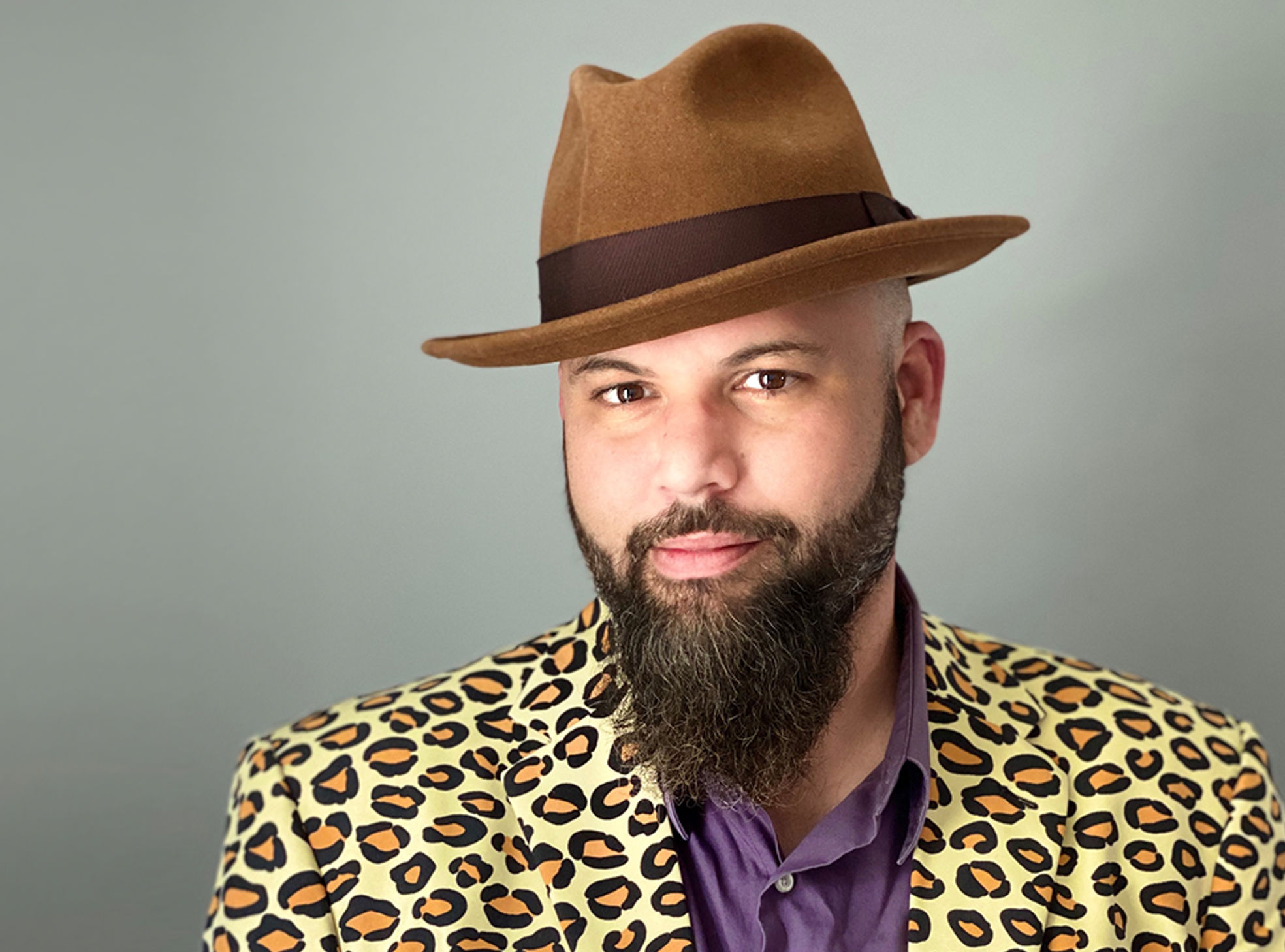 This is a photo of Bryan Dean. He has a dark brown beard and mustache and is wearing a brown hat and cheetah print blazer.