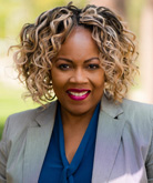 Woman with curly blonde and dark brown hair wearing gray blazer and blue shirt. 