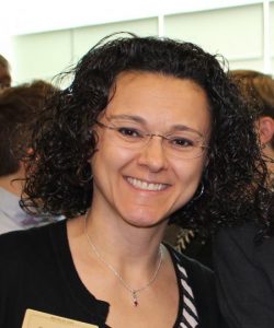 Image is a portrait photo of a smiling Latina woman, mid-40's, with shoulder length dark brown curly hair. Woman wears frameless oval glasses, a black cardigan, and a black-and-white striped shirt. 