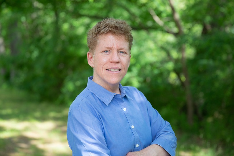 Image of Dr. Kristen Renn. Caucasian woman with short light brown hair facing the camera in a blue shirt. Background is of blurry green foliage. 