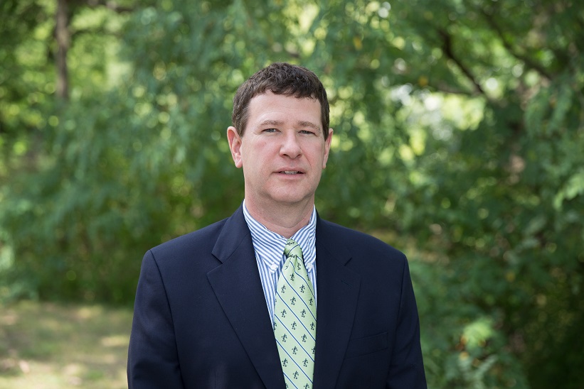 Image of Dr. Matt Wawrzynski. Caucasian man with short brown hair facing the camera wearing a dark blue suit jacket, striped white and blue collared shirt, and light green tie. Background is of blurry green foliage. 