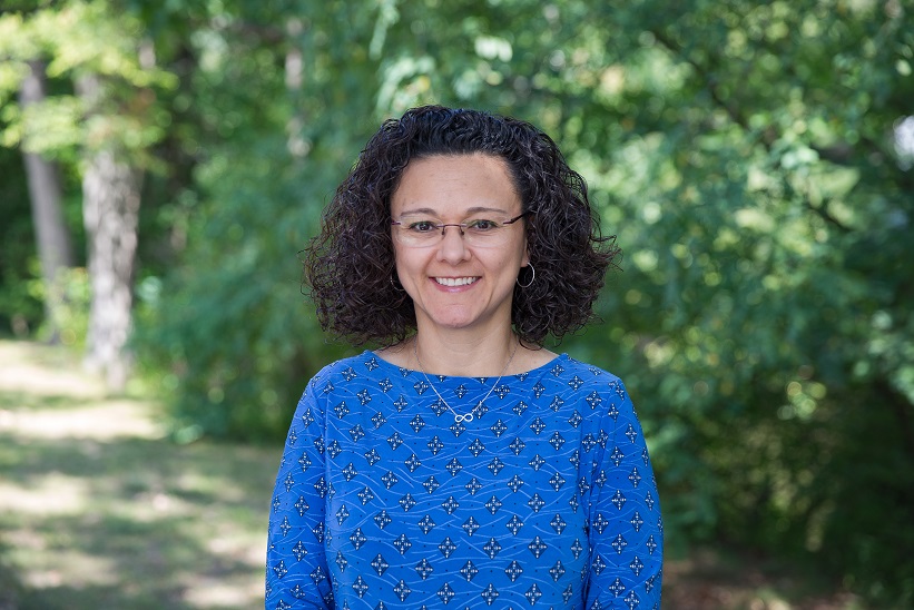 Image of Dr. Patricia Marin. Latina woman with dark curly hair facing the camera, wearing a blue shirt, and also wearing glasses. Background is of blurry green foliage. 