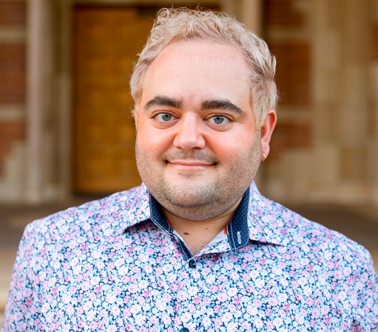 Image of Dr. Justin Gutzwa, a caucasian person wearing a floral patterned blue and pink collared button down shirt. The background is blurred and features brick walls.