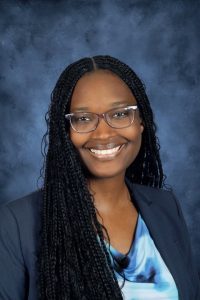 Image of a smiling young Black woman from the shoulders up. The woman wears a navy blue blazer and a blue and white top. The woman also wears light purple glasses and has long micro-braided hair. The woman sits in front of a mottled dark blue professional photo background.