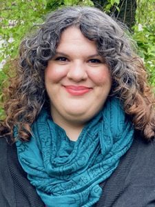 Image is a portrait-style photo of a smiling woman, mid-30's, with olive skin, gray and brown curly hair, and brown eyes. Woman wears an emerald green scarf and a black cardigan. Woman stands in front of a leafy background. 