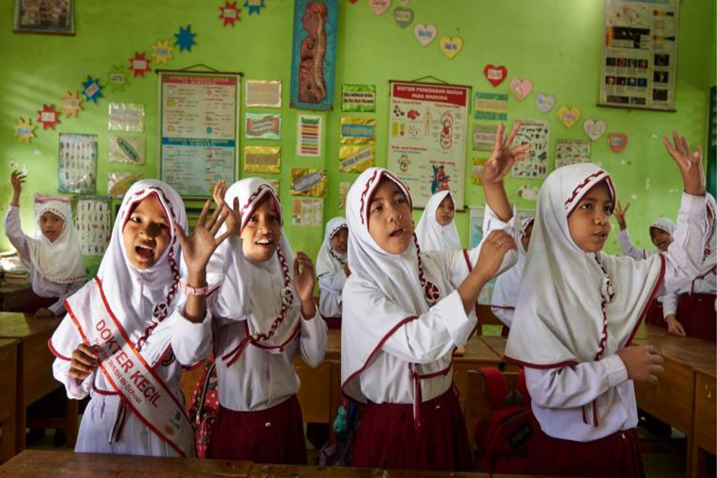 Four girls raise their hands while singing and dancing in a classroom. They wear white tops and white head coverings with red fringes. Other classmates are behind them in a classroom painted bright green and covered in artwork and miscellaneous signage.
