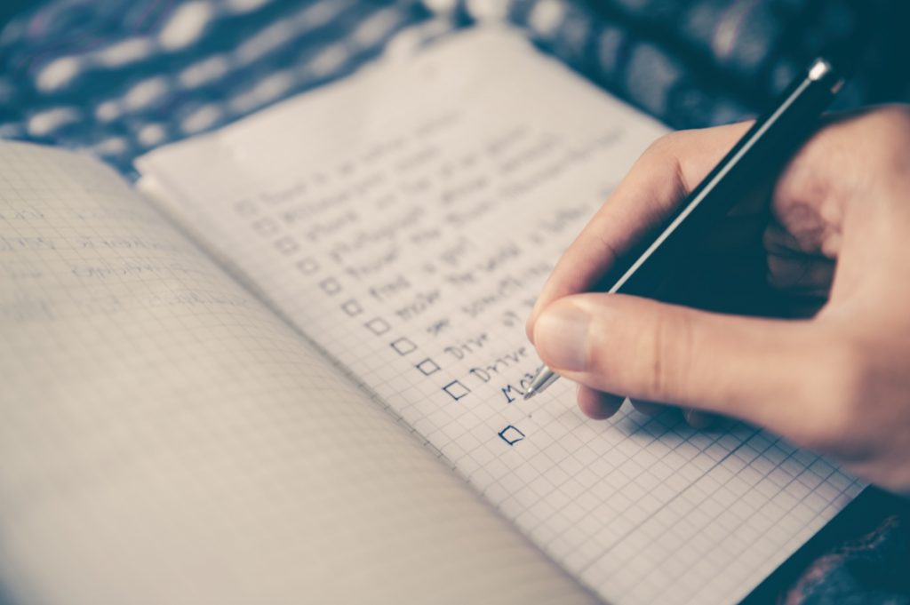 This is a picture of someone making a checklist in a notebook. A hand holding a pen appears in the image with the notebook.
