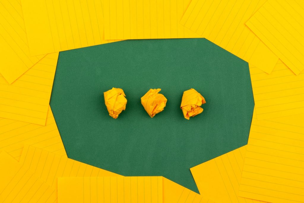 This is a picture of yellow notecards scattered around to create an outline of a a dialogue bubble in the center. The green dialogue bubble has 3 crumpled notecards in the center that appear as dot, dot, dot to indicate that someone is thinking or typing.
