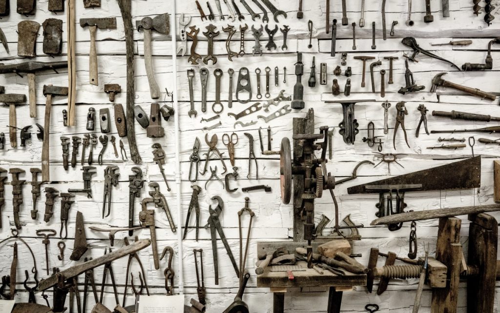 This is a picture of a white wall that has at least 50 wooden and metal tools displayed on it. There is a large industrial machine in the photo as well. 