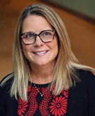 Julie Helber headshot; she wears a red and black floral top with a black cardigan over it. Her blonde hair is about shoulder-length. She wears dark-rimmed glasses.