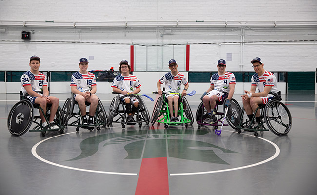 Six memebrs of the MSU wheelchair floorball team sit in wheelchairs and smile toward the camera. They are on the floor of a gym with a Spartan helmet directly in the center of the floor. 