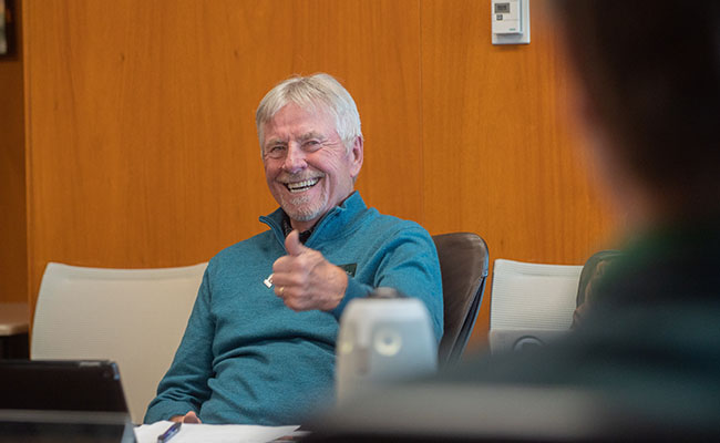 Bob Chapla grins during an Alumni Board meeting. He has his thumb up in the air in approval. He wears a long-sleeve blue shirt. 