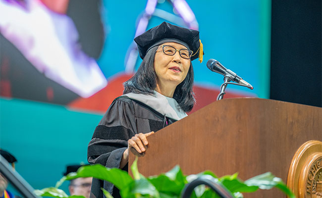 Okhee Lee stands delivers a speech at podium dressed in full commencement regalia. 