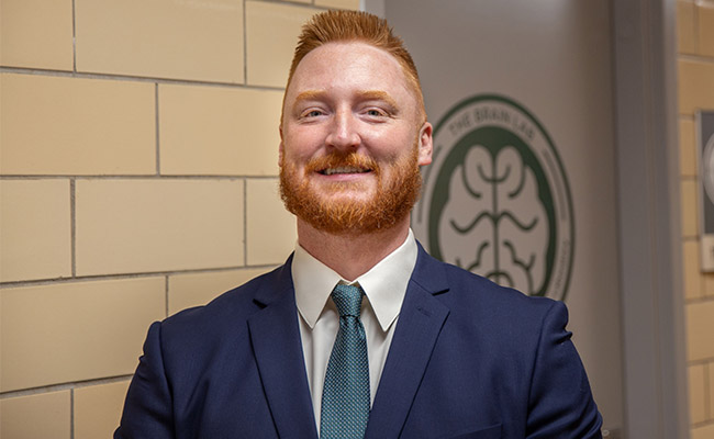 Aaron stands outside of The Brain Lab at MSU. He wears a navy blazer over a white button down and blue tie. He has red hair cropped short and a beard.