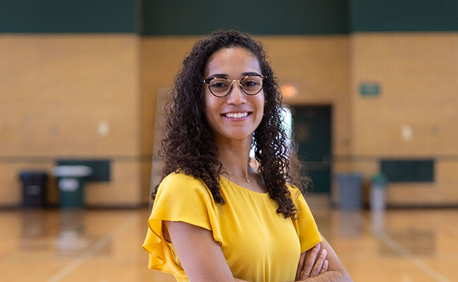 Brooks stands in a gym, smiling at the camera with her arms crossed. She wears a yellow blouse. Her dark hair is long and flows about her shoulders. She wears dark-rimmed glasses.