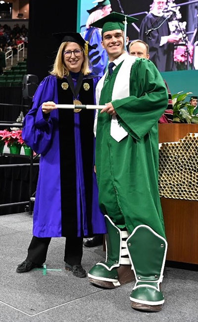 Rau standing on a stage with Interim President Woodruff while holding his diploma. Rau is wearing academic regalia and Sparty Boots. Both Rau and Interim President Woodruff are smiling. 