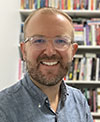 Adam Greteman smiling in front of a shelf of books. Greteman is wearing a blue shirt and glasses. 