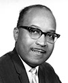 Black and white photo of James McCray. He is wearing a dark suit jacket, light colored shirt and a light colored tie. He also is wearing glasses and smiling. 