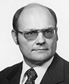 Black and white photo of John Haubenstricker. He is wearing a dark suit jacket, a light colored shirt and a dark tie with dots. Haubenstricker is also wearing glasses. 