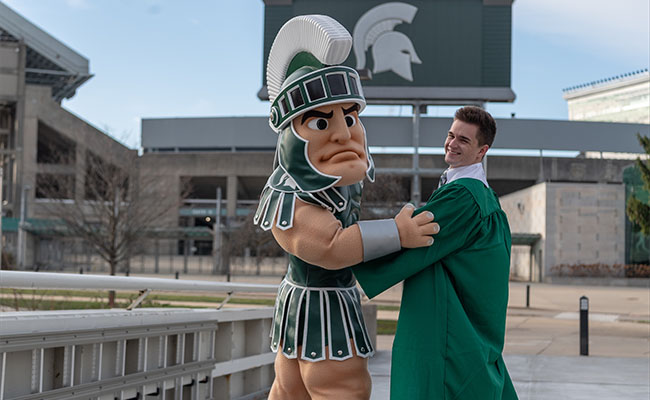 Rau posing with Sparty in academic regalia in front of Spartan Stadium. 