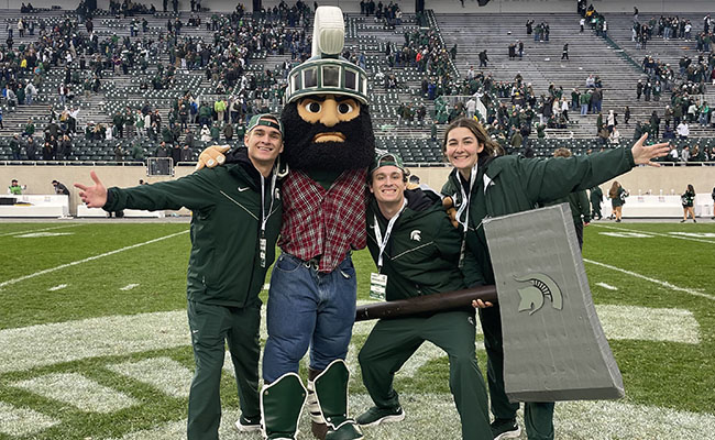 Rau and two others posing with Sparty dressed as a lumberjack in Spartan Stadium. 

