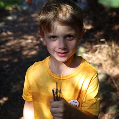 A child wearing a yellow shirt, smiling at the camera. He holding several small twigs in his fist. 