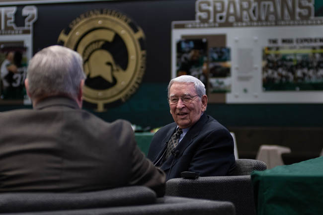 Two people sitting in conversation with a Spartan logo visible in the background.