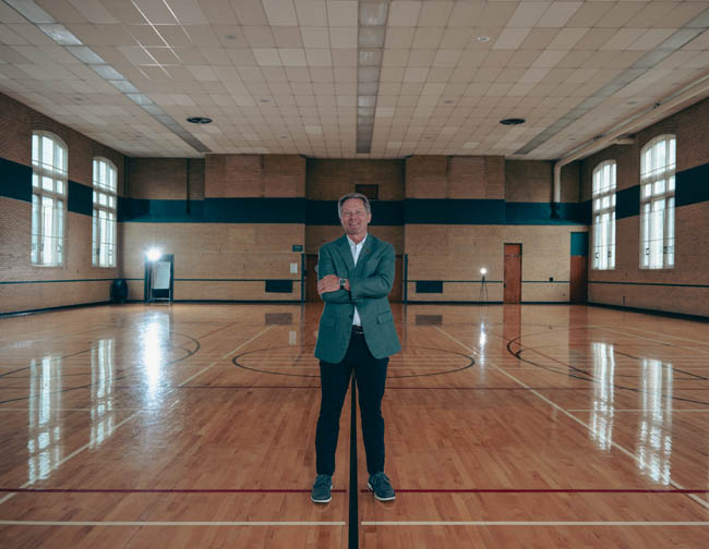 A man standing in an empty gymnasium wearing a green suit.