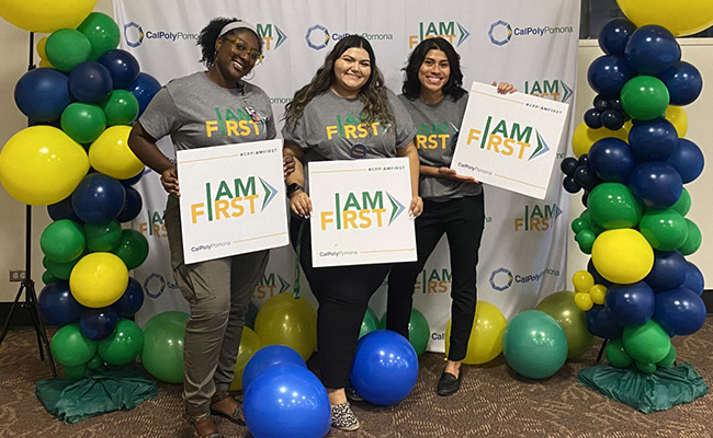 Three smiling individuals holding 'I AM FIRST' signs in front of a backdrop featuring the Cal Poly Pomona logo. They are surrounded by colorful balloon decorations in green, yellow, and blue, celebrating a first-generation student event.