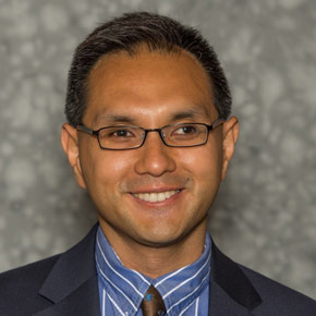 Peter De Costa smiles toward the camera. The background is a blurred gray. De Costa wears a blue and white stripped shirt and a dark tie along with a dark blazer. He wears glasses with dark rims. 