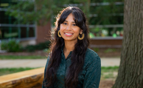 Tasminda Dhaliwal sits on a bench outside of Erickson Hall. She wears a dark green button-down shirt, and big hoop earings in a golden hue. Her long brown hair is half pulled up. 