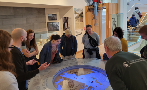 Students are gathered around a circular table in the Galway City Museum, leaning in toward an exhibit about the topography of the town. In the forefront of the photo, one person wears a College of Education shirt. 