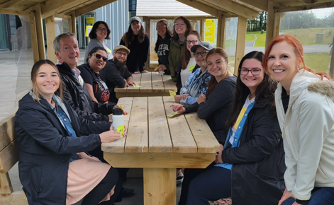 12 MAET students sit at picnic tables and smile toward the camera. Program director Liz Owens Bolts stands to the right, smiling. 