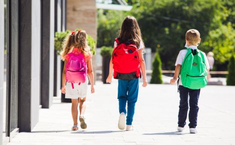 Three elementary-aged students walk outside of a school with their backs towards the camera. 