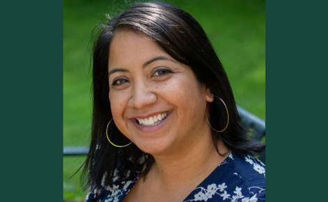 Headshot of Rodríguez smiling at the camera while sitting on a bench. She wears a navy blouse with white flowers on it, and gold hoop earrings. Her dark hair falls about shoulder-length. 